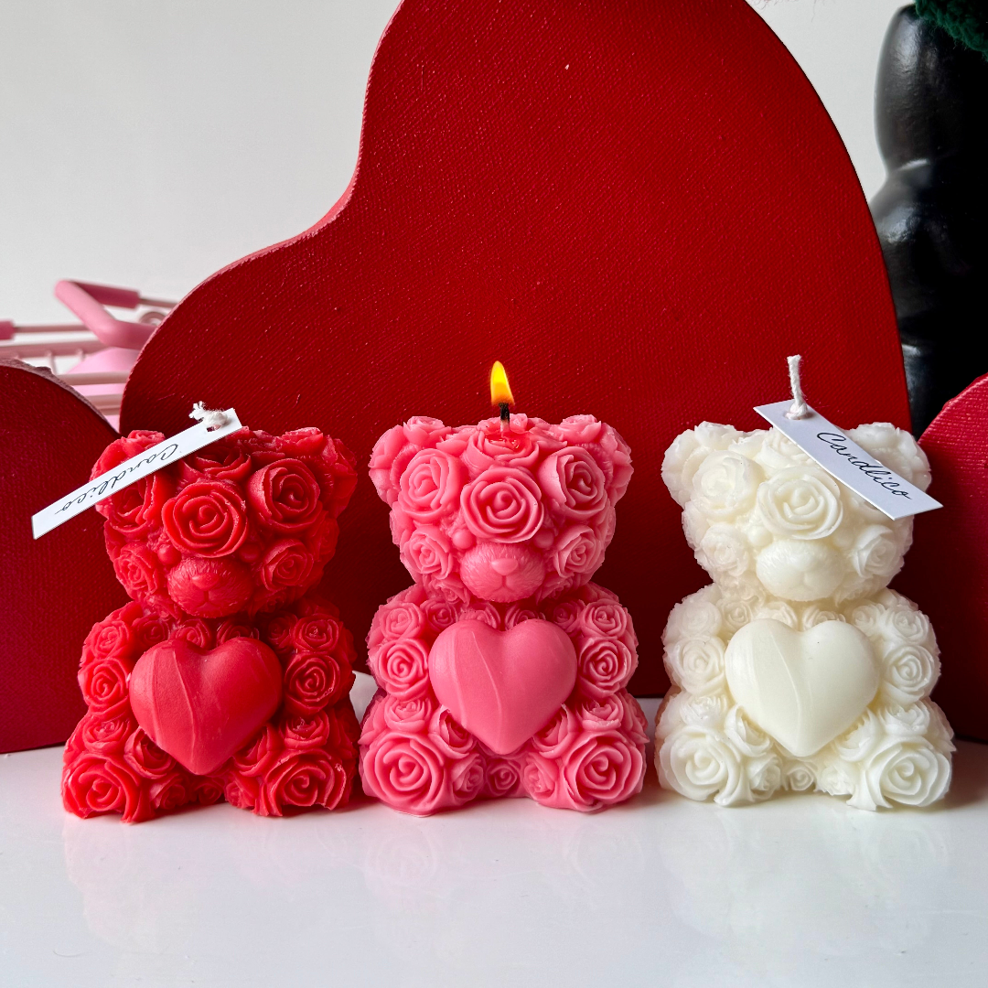 Three teddy bear-shaped candles in red, pink, and white with heart designs against a red heart-shaped backdrop.